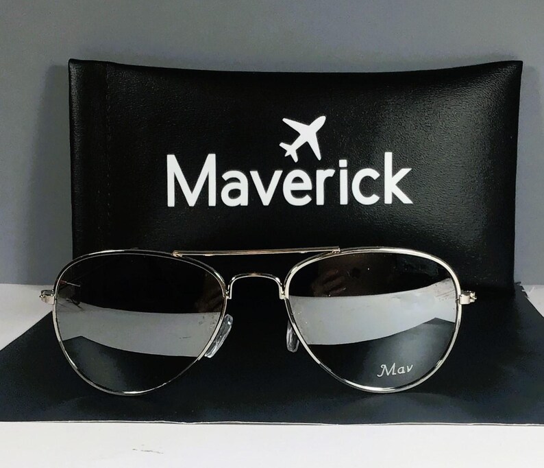 May include: Aviator sunglasses with thin silver metal frames and reflective silver lenses. The right lens features the text Mav in white. Behind the sunglasses is a black rectangular case with the word Maverick and an airplane icon.