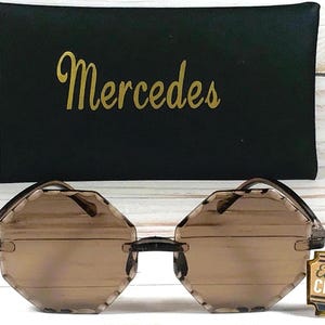 May include: Octagon-shaped sunglasses with brown lenses and a black frame. The sunglasses are on a white surface with a black fabric case that says "Mercedes" in gold lettering. A small sign with the text "Eat Cake & CELEBRATE" is in the lower right corner of the image.