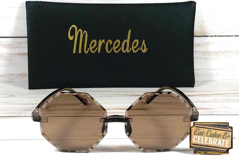May include: Octagon-shaped sunglasses with brown lenses and a black frame. The sunglasses are on a white surface with a black fabric case that says "Mercedes" in gold lettering. A small sign with the text "Eat Cake & CELEBRATE" is in the lower right corner of the image.