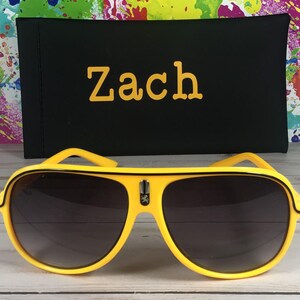 Puede incluir: Gafas de sol de aviador amarillas con lentes oscuras y un estuche negro. El estuche tiene el nombre "Zach" en letras amarillas. El fondo presenta un dise&ntilde;o de salpicaduras de pintura coloridas.