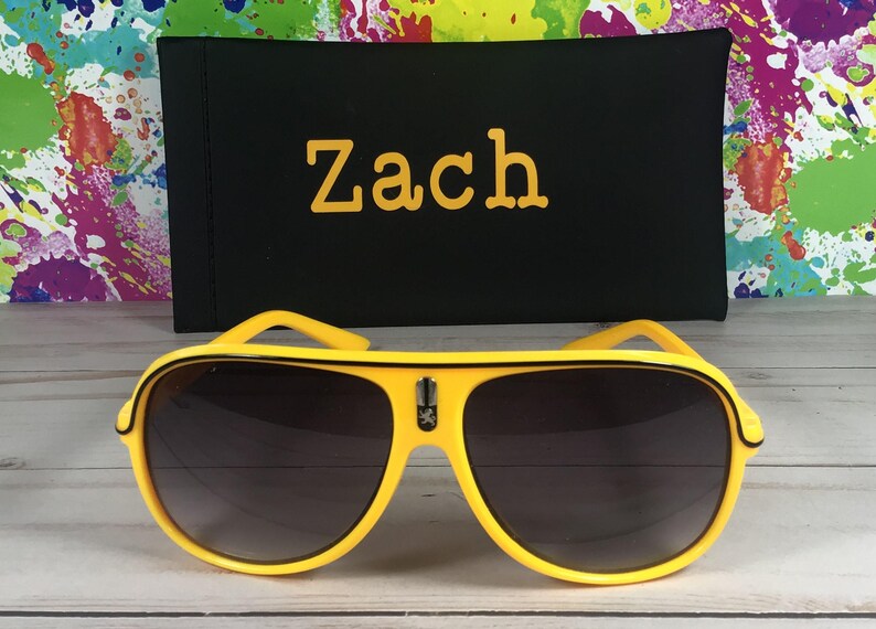 Puede incluir: Gafas de sol de aviador amarillas con lentes oscuras y un estuche negro. El estuche tiene el nombre "Zach" en letras amarillas. El fondo presenta un dise&ntilde;o de salpicaduras de pintura coloridas.
