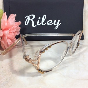 Puede incluir: Gafas transparentes con detalles dorados y peque&ntilde;os tachuelas decorativas. Las gafas est&aacute;n delante de un estuche negro con el nombre "Riley" en escritura blanca. Flores rosas en el fondo.