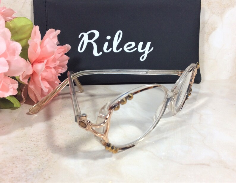 Puede incluir: Gafas transparentes con detalles dorados y peque&ntilde;os tachuelas decorativas. Las gafas est&aacute;n delante de un estuche negro con el nombre "Riley" en escritura blanca. Flores rosas en el fondo.