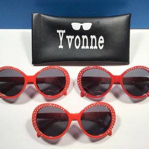 May include: Three pairs of red oval sunglasses with rhinestones around the frames. The sunglasses have dark lenses. The sunglasses are on a white surface.