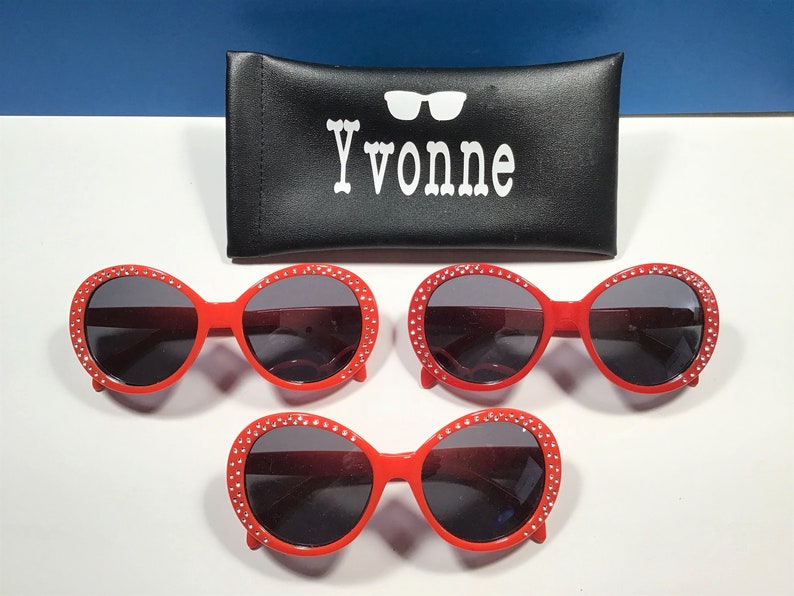 May include: Three pairs of red oval sunglasses with rhinestones around the frames. The sunglasses have dark lenses. The sunglasses are on a white surface.