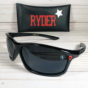 Op de afbeelding: Zwarte zonnebril met een rood en blauw logo aan de zijkant. De zonnebril wordt geleverd met een zwarte etui met het woord "RYDER" in rode letters en een witte ster.