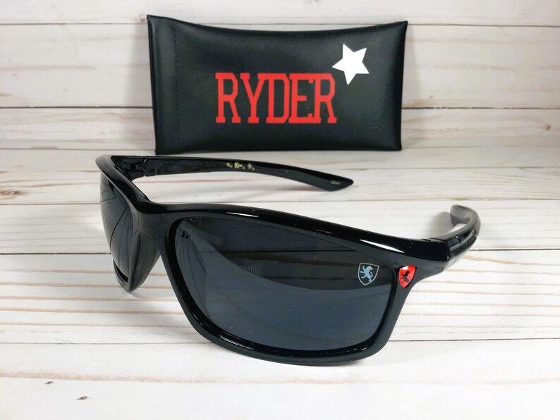 Op de afbeelding: Zwarte zonnebril met een rood en blauw logo aan de zijkant. De zonnebril wordt geleverd met een zwarte etui met het woord "RYDER" in rode letters en een witte ster.