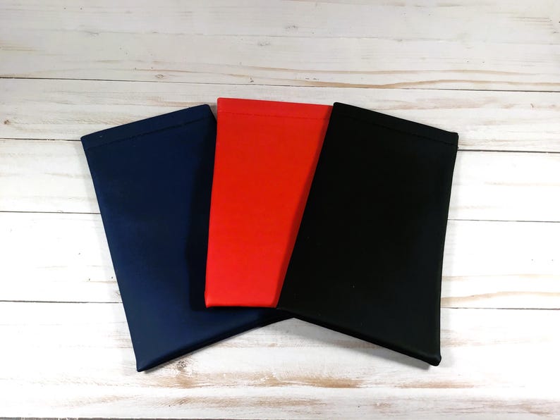 Puede incluir: Tres estuches para gafas en azul marino, rojo y negro. Los estuches son rectangulares y parecen estar hechos de un material suave y liso. Est&aacute;n colocados sobre una superficie de madera blanca.