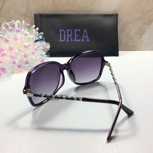 May include: Purple sunglasses with a silver chain detail on the arms. The sunglasses are in a black case with the word "DREA" printed on it.