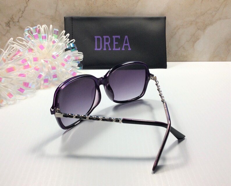 May include: Purple sunglasses with a silver chain detail on the arms. The sunglasses are in a black case with the word "DREA" printed on it.