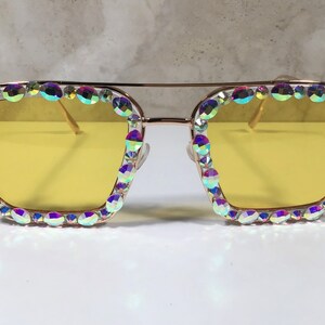 Peut inclure: Lunettes de soleil &agrave; monture dor&eacute;e avec des verres jaunes et des strass iris&eacute;s autour de la monture.