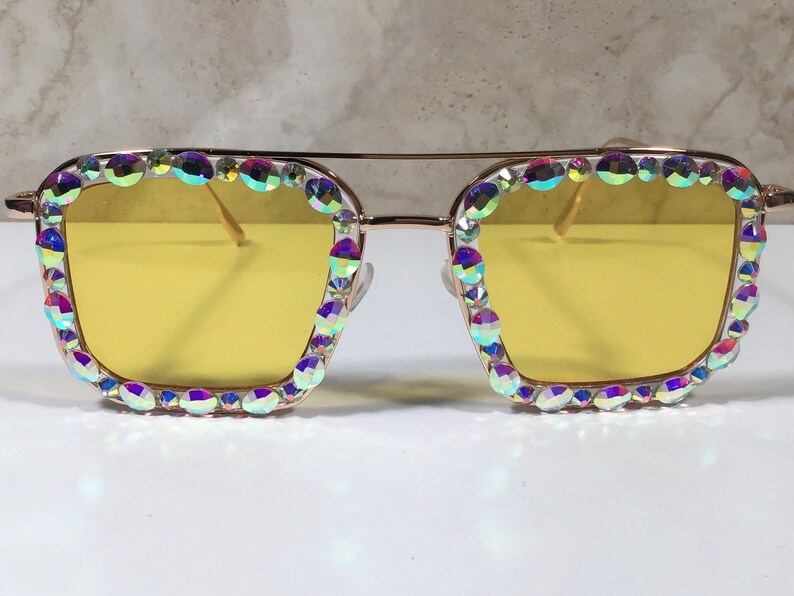 Peut inclure: Lunettes de soleil &agrave; monture dor&eacute;e avec des verres jaunes et des strass iris&eacute;s autour de la monture.