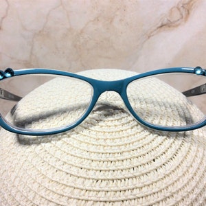 May include: Teal-colored eyeglasses with clear lenses. The frames are adorned with small, round, blue gemstones at the outer corners. The glasses rest on a white, woven hat, set against a neutral background.