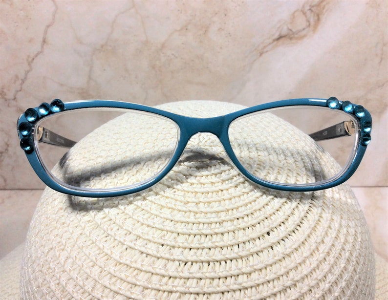 May include: Teal-colored eyeglasses with clear lenses. The frames are adorned with small, round, blue gemstones at the outer corners. The glasses rest on a white, woven hat, set against a neutral background.