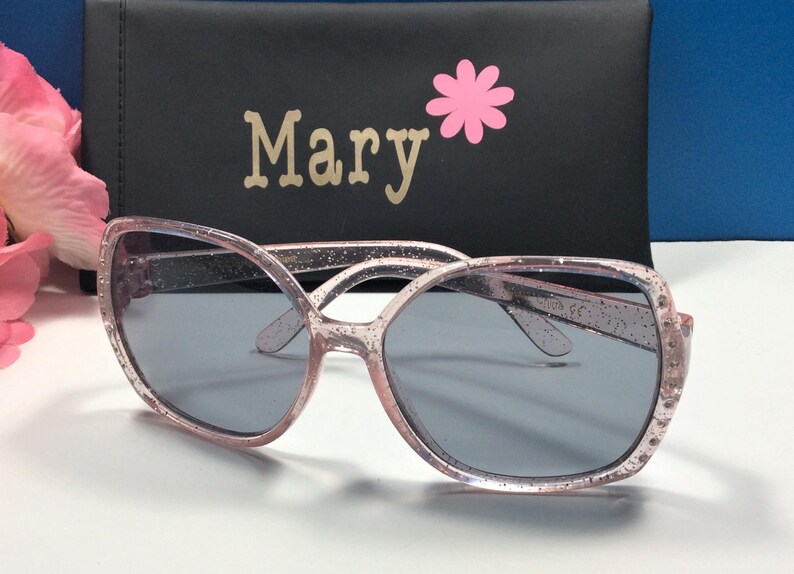 May include: Pink glitter sunglasses with a black case that says "Mary" on it with a pink flower.