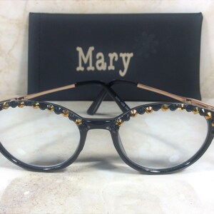May include: Black cat-eye glasses with gold accents and decorative jewels along the top rim. The glasses have clear lenses and a gold-coloured frame. A black case with the name "Mary" is in the background.