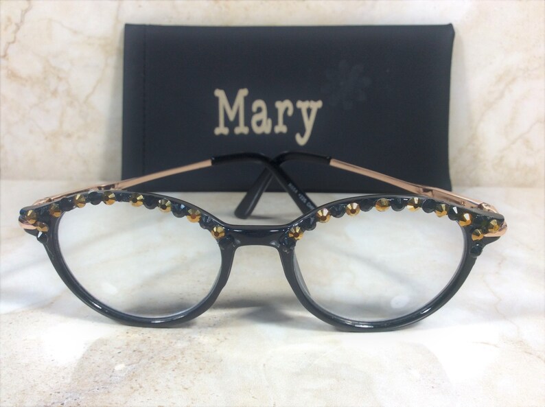 May include: Black cat-eye glasses with gold accents and decorative jewels along the top rim. The glasses have clear lenses and a gold-coloured frame. A black case with the name "Mary" is in the background.