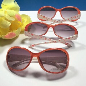 May include: Three pairs of orange sunglasses with white frames and pink hearts printed on the arms. The sunglasses have a dark grey lens. The bottom pair has the name "Samantha" printed on the right lens.