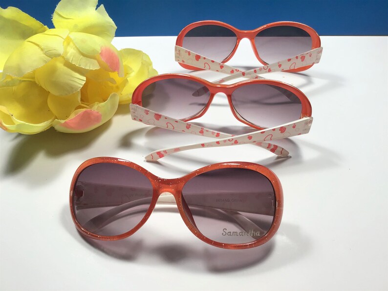 May include: Three pairs of orange sunglasses with white frames and pink hearts printed on the arms. The sunglasses have a dark grey lens. The bottom pair has the name "Samantha" printed on the right lens.