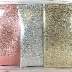 May include: Three rectangular eyeglass cases in a row. The cases are in shades of pink, silver, and gold. Each case has a textured, dotted pattern. The cases are made of a shiny material and are standing upright.