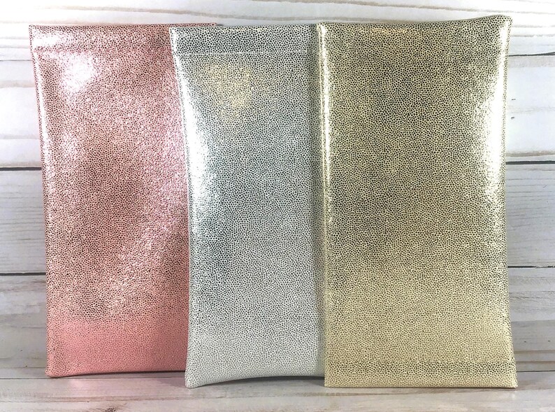 May include: Three rectangular eyeglass cases in a row. The cases are in shades of pink, silver, and gold. Each case has a textured, dotted pattern. The cases are made of a shiny material and are standing upright.