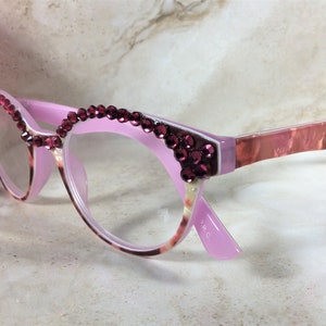 K&ouml;nnte beinhalten: Rosa Cat-Eye-Brille mit burgunderfarbenen Strasssteinen am oberen Rand und an den Ecken. Die B&uuml;gel haben ein marmoriertes rosa-braunes Muster. Die Gl&auml;ser sind klar. Eine rosa Blume ist im Hintergrund.
