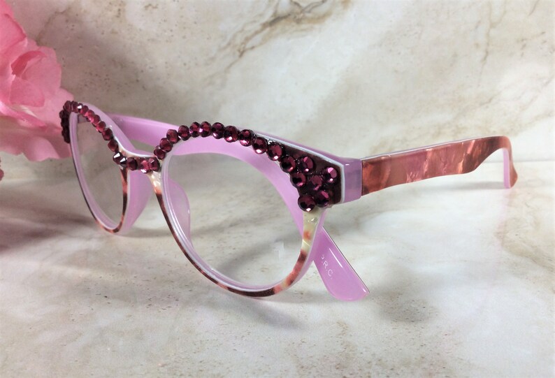 K&ouml;nnte beinhalten: Rosa Cat-Eye-Brille mit burgunderfarbenen Strasssteinen am oberen Rand und an den Ecken. Die B&uuml;gel haben ein marmoriertes rosa-braunes Muster. Die Gl&auml;ser sind klar. Eine rosa Blume ist im Hintergrund.