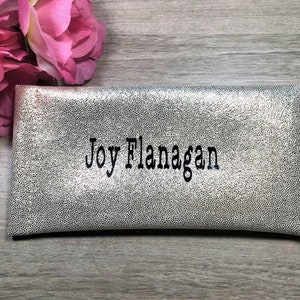 Pu&ograve; includere: Pochette argentata con un motivo a pois strutturato. Il nome "Joy Flanagan" &egrave; ricamato in nero. Fiori rosa sono visibili sullo sfondo. La pochette &egrave; rettangolare e sembra essere realizzata in un materiale morbido.