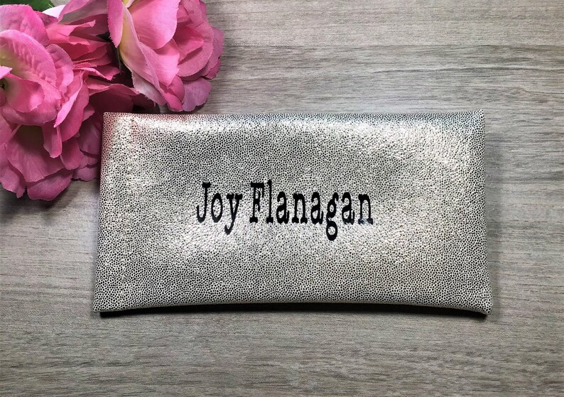 Pu&ograve; includere: Pochette argentata con un motivo a pois strutturato. Il nome "Joy Flanagan" &egrave; ricamato in nero. Fiori rosa sono visibili sullo sfondo. La pochette &egrave; rettangolare e sembra essere realizzata in un materiale morbido.