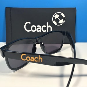 May include: Black sunglasses with a Coach logo on the side and a black case with a soccer ball and the word "Coach" on it.