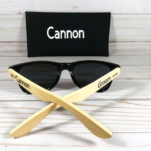 May include: Black sunglasses with bamboo arms. The arms are engraved with the words "Cannon" and "Groom". The sunglasses come with a black carrying case that has the word "Cannon" embroidered on it.