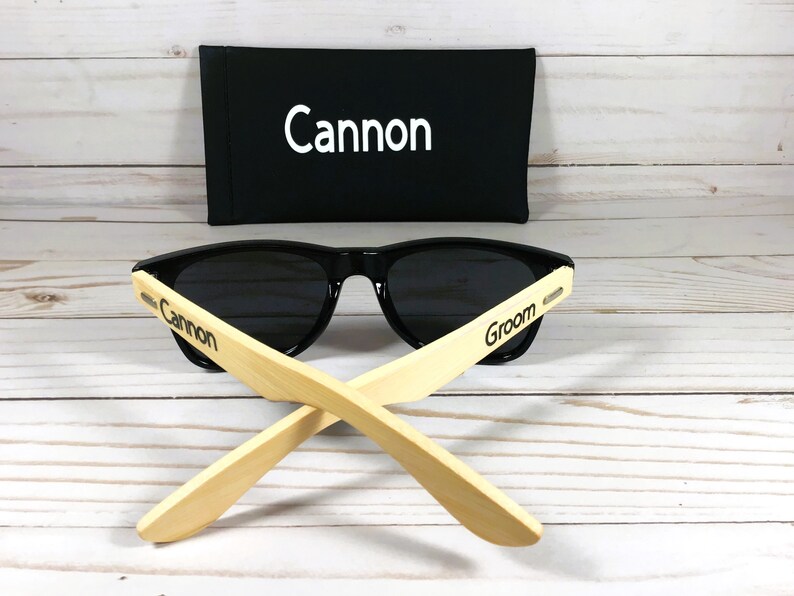 May include: Black sunglasses with bamboo arms. The arms are engraved with the words "Cannon" and "Groom". The sunglasses come with a black carrying case that has the word "Cannon" embroidered on it.