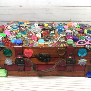 May include: A wooden treasure chest, adorned with a multitude of colorful embellishments. The top is covered in various trinkets, including beads, gems, and small plastic figures. The chest features a red interior, a leather handle, and decorative gems on the front.