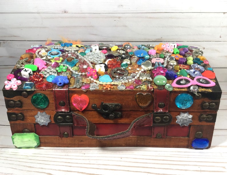 May include: A wooden treasure chest, adorned with a multitude of colorful embellishments. The top is covered in various trinkets, including beads, gems, and small plastic figures. The chest features a red interior, a leather handle, and decorative gems on the front.