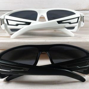 Pu&ograve; includere: Sono mostrati due paia di occhiali da sole, uno bianco e uno nero. Il paio bianco, in alto, ha un design moderno con montature angolari e lenti scure. Gli occhiali da sole neri sottostanti rispecchiano il design. Entrambi i paia hanno aste curve.