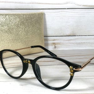 May include: Black round-frame glasses with thin gold-tone metal temples and small gold-tone studs on the outer corners of the frames. The glasses feature clear lenses and are positioned in front of a textured gold-colored rectangular case.
