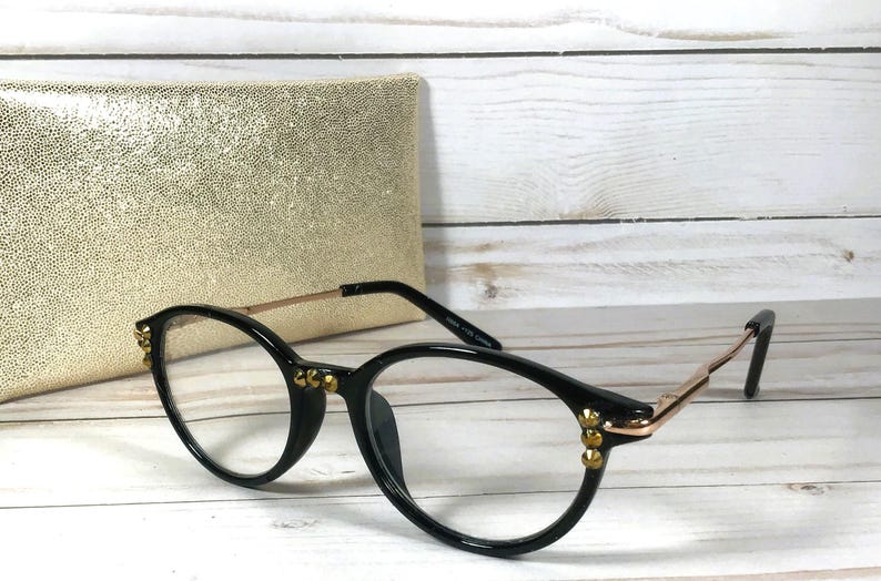 May include: Black round-frame glasses with thin gold-tone metal temples and small gold-tone studs on the outer corners of the frames. The glasses feature clear lenses and are positioned in front of a textured gold-colored rectangular case.