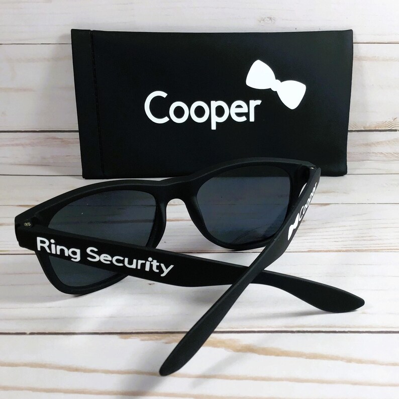Op de afbeelding: Zwarte zonnebril met donkere glazen en de witte tekst Ring Security op de pootjes. Achter de zonnebril staat een zwart rechthoekig etui met de witte tekst Cooper en een witte strik als afbeelding op de voorzijde van het etui.