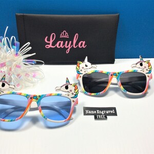 May include: Two pairs of colorful unicorn sunglasses with rainbow stripes and a unicorn head on each side. The sunglasses have blue and gray lenses. The sunglasses are on a white surface. The text "Name Engraved FREE" is visible in the image.