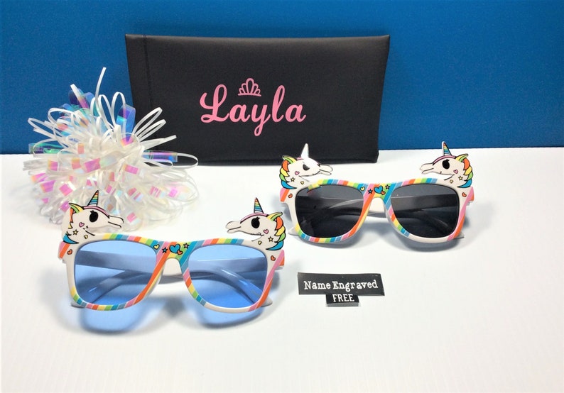 May include: Two pairs of colorful unicorn sunglasses with rainbow stripes and a unicorn head on each side. The sunglasses have blue and gray lenses. The sunglasses are on a white surface. The text "Name Engraved FREE" is visible in the image.