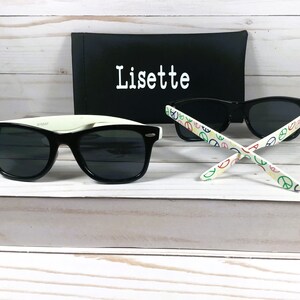 Op de afbeelding: Zwarte zonnebril met een wit montuur en peace-sign patroon op de armen. De zonnebril zit in een zwarte etui met de naam "Lisette" erop.