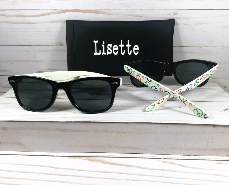 Op de afbeelding: Zwarte zonnebril met een wit montuur en peace-sign patroon op de armen. De zonnebril zit in een zwarte etui met de naam "Lisette" erop.