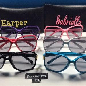 May include: Six pairs of sunglasses with different colored frames, including purple, pink, red, black, white, and blue. The sunglasses have a cat-eye shape and are arranged in two rows of three. The sunglasses are on a white surface with a black sign that says "Name Engraved FREE".