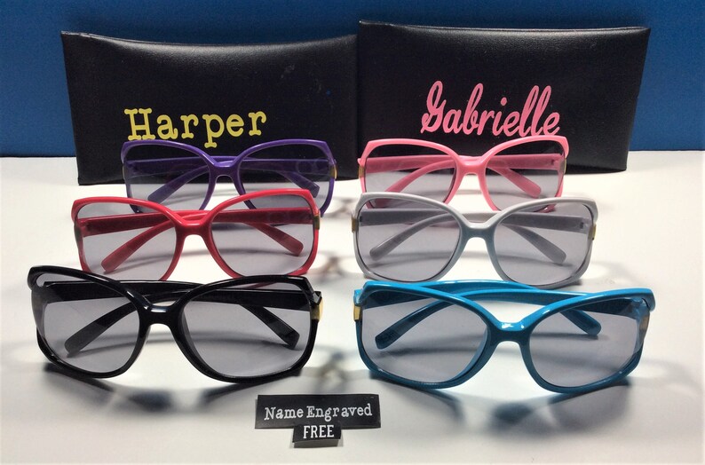 May include: Six pairs of sunglasses with different colored frames, including purple, pink, red, black, white, and blue. The sunglasses have a cat-eye shape and are arranged in two rows of three. The sunglasses are on a white surface with a black sign that says "Name Engraved FREE".