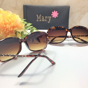May include: Tortoise shell sunglasses with brown lenses and a gold chain detail. The sunglasses are folded in half and the case is visible in the background. The case is black with the word "Mary" and a pink flower.