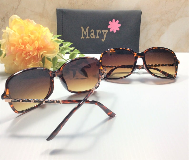 May include: Tortoise shell sunglasses with brown lenses and a gold chain detail. The sunglasses are folded in half and the case is visible in the background. The case is black with the word "Mary" and a pink flower.