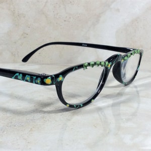 May include: Black eyeglasses with a floral design in teal and yellow. The frames have a decorative row of green rhinestones along the top edge. The text "China" is printed on the temple.