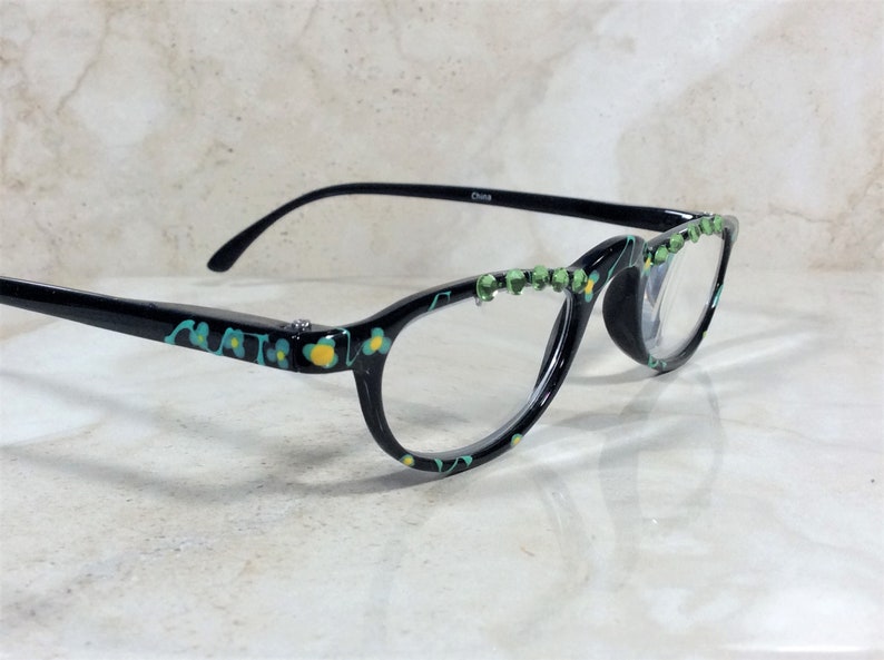 May include: Black eyeglasses with a floral design in teal and yellow. The frames have a decorative row of green rhinestones along the top edge. The text "China" is printed on the temple.