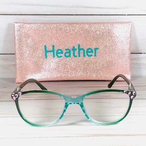 May include: A pair of cat-eye glasses with a translucent green frame and small pink rhinestones at the outer corners, positioned in front of a rectangular rose gold glitter case with the name Heather printed in teal blue lettering.