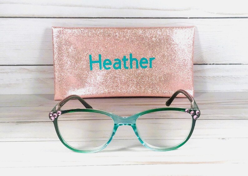 May include: A pair of cat-eye glasses with a translucent green frame and small pink rhinestones at the outer corners, positioned in front of a rectangular rose gold glitter case with the name Heather printed in teal blue lettering.